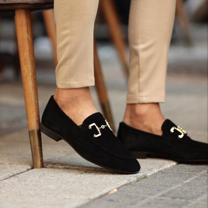 Black suede loafers with polished gold buckle trim, worn with beige chinos