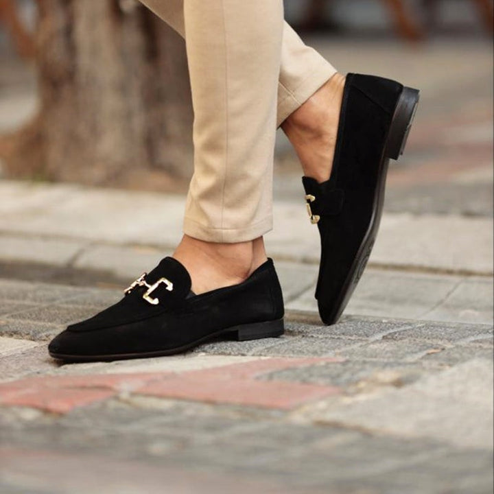 Man crossing legs in black suede loafers with gold buckle accent, outdoor setting