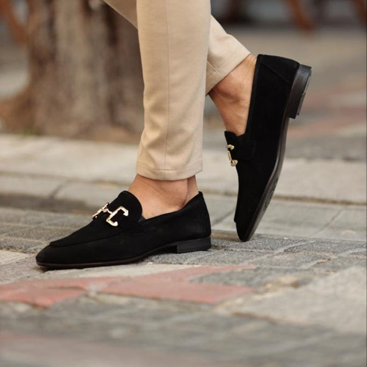 Black suede loafers featuring gold buckle detail, styled with beige trousers