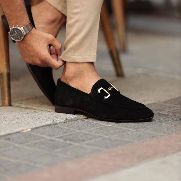 Black suede slip-on loafers with gold trim, worn beneath tailored chinos