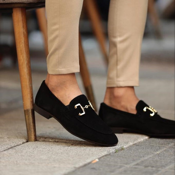 Black suede loafers with gold buckle accent, paired with beige tailored trousers