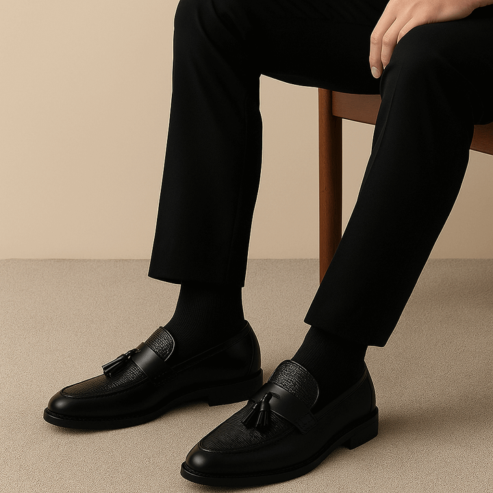 Man wearing black leather tassel loafers with tailored black trousers, seated pose
