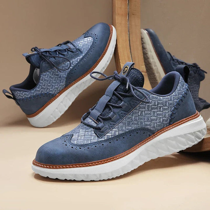 Pair of navy woven leather brogue trainers displayed against a wooden stand