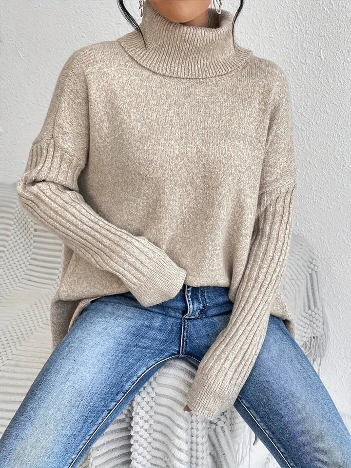 Seated model wearing beige oversized turtleneck jumper with ribbed sleeves