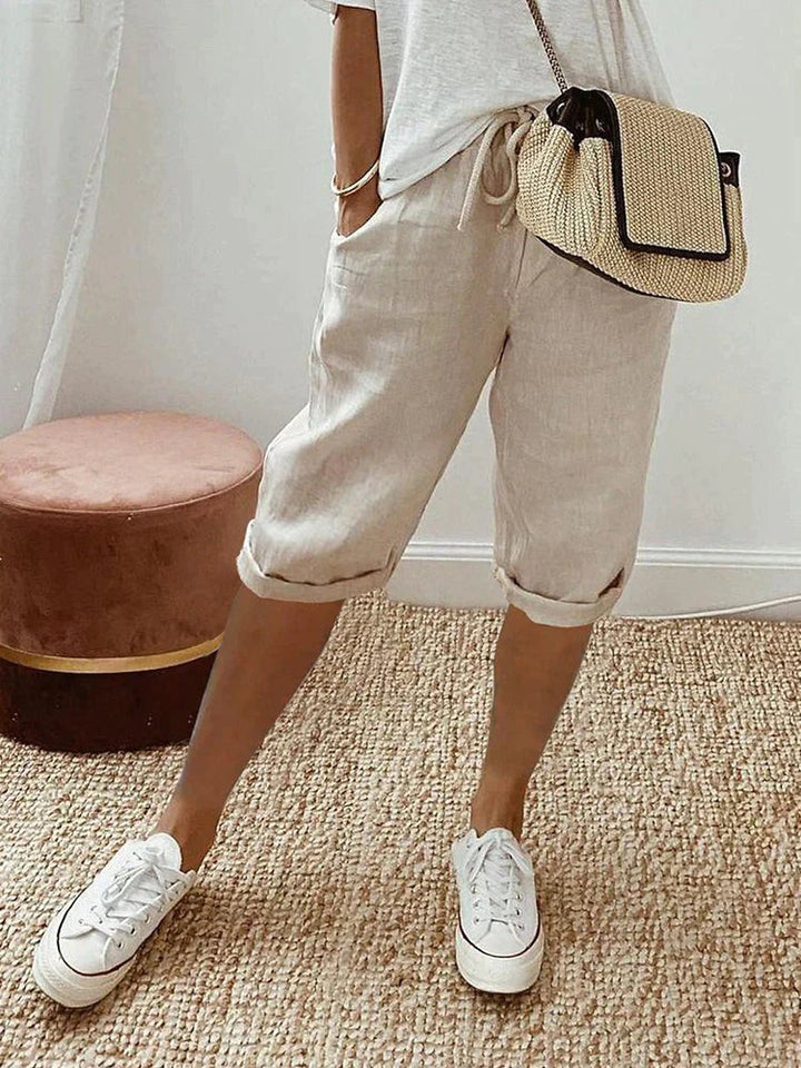 Beige linen turn-up trousers styled with white trainers and a woven crossbody bag