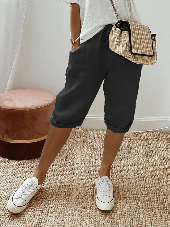 Black linen turn-up trousers paired with a straw crossbody bag and white trainers
