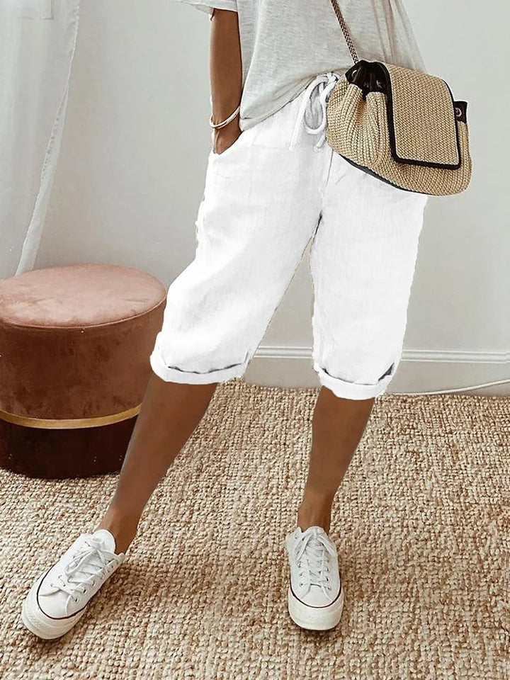 White linen turn-up trousers styled casually with white trainers and a woven bag