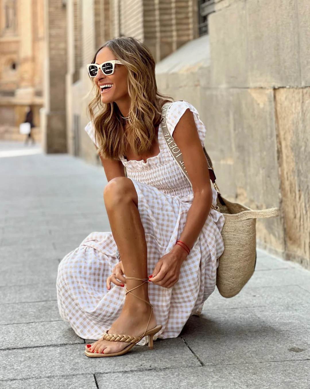 Model sitting on pavement wearing beige gingham smocked maxi dress with tie sandals