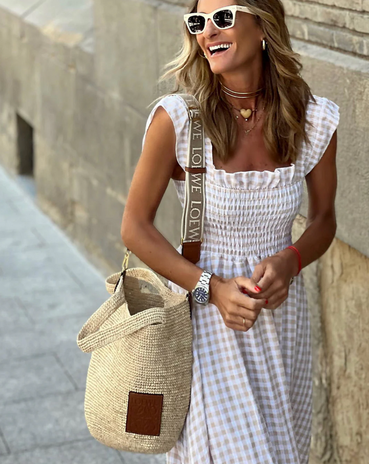 Smocked bodice detail on beige gingham maxi dress paired with woven shoulder bag