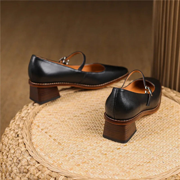 Black leather Mary Jane shoes with block heels shown from behind on straw surface