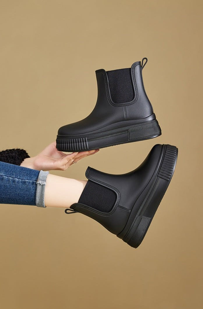 Black Chelsea rain boots held in hand beside model wearing matching pair
