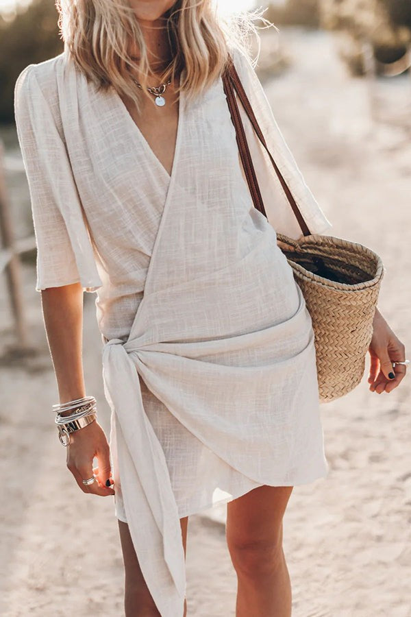 Cream linen wrap mini dress with tie waist and V-neckline, worn on a beach